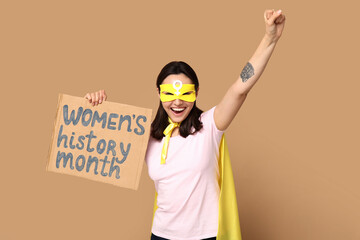Young woman in superhero costume holding cardboard with text WOMEN'S HISTORY MONTH on beige...