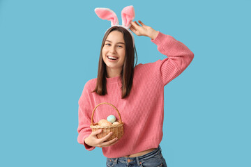 Pretty young woman with bunny ears and basket of Easter eggs on blue background