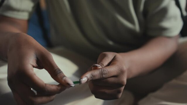Close-up shot of hands of unrecognizable African American or Asian woman with diabetes sitting on bed, pricking finger with lancet, smearing blood onto test strip and inserting it into meter