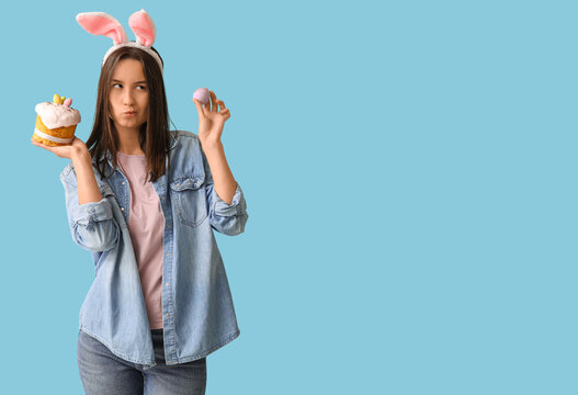 Thoughtful young woman with bunny ears, Easter cake and egg on blue background
