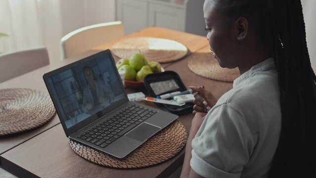 Medium shot of African American woman with diabetes sitting in front of laptop, having telemedicine appointment with GP on video call, reporting on her condition, showing glucose monitor device on arm