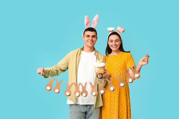 Happy young couple with Easter bunny ears, cake and garland on blue background