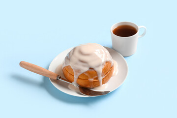 Plate with tasty glazed cinnamon bun, fork and cup of tea on blue background
