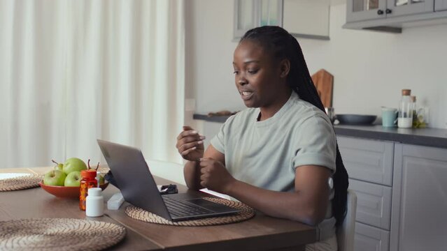 Medium shot of smiling African American woman with diabetes sitting at kitchen table in front of laptop, having video call with friend, chatting and demonstrating new glucose monitoring device on arm