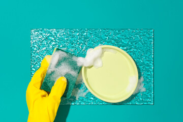 Hand wearing yellow rubber gloves holds a sponge, white foam and a ceramic plate on a glass sheet on a turquoise background. Creative space for advertising.