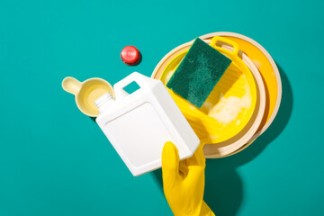 A hand wearing a yellow rubber glove holds a bottle of detergent and pours it into a water scoop, next to a sponge with dirty dishes. Copy space for advertising.