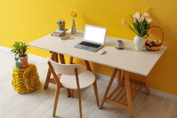 Workplace with laptop, Easter eggs and flowers near yellow wall in office