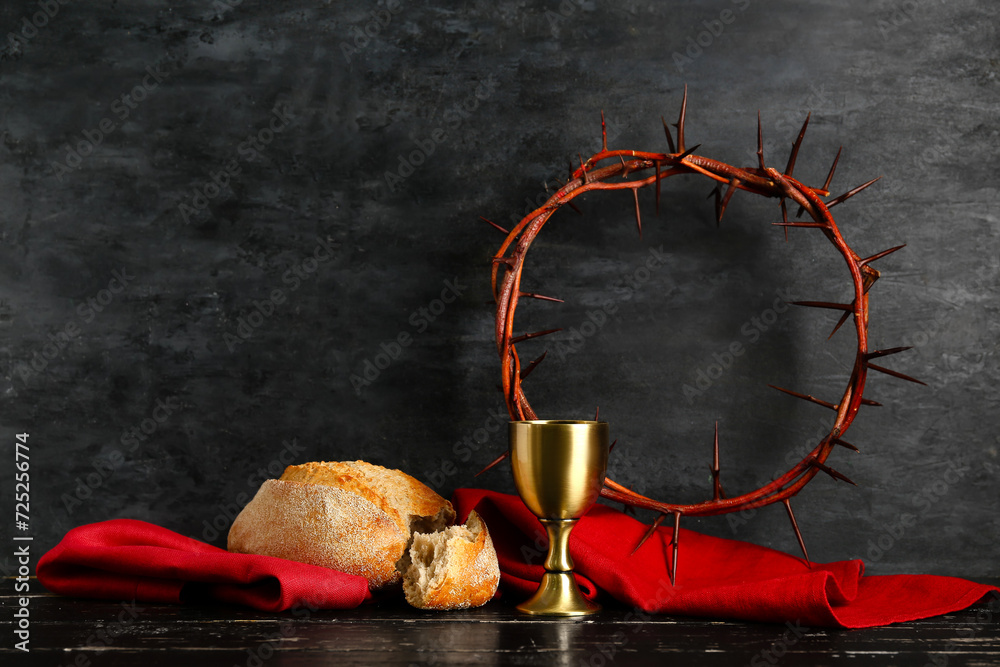 Wall mural crown of thorns with chalice, red cloth and bread on black wooden table against dark background