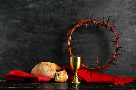 Crown of thorns with chalice, red cloth and bread on black wooden table against dark background