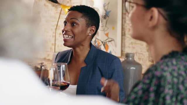 black businesswoman in formal clothes sitting in restaurant and talking emotionally, colleagues on foreground, wine glasses on table. Business partners negotiating during lunch