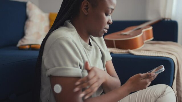 Medium close-up side shot of young African American woman with diabetes sitting on floor by couch in living room, touching glucose monitoring device on arm, then saving data on smartphone app