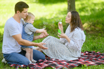 Fototapeta premium Cute young family sits on a blanket in park, mother blows soap bubbles, father and child catch them, play, have fun togetheron a warm sunny weekend.