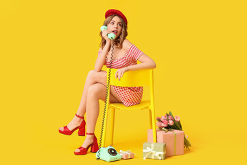 Young woman talking by retro telephone with gift boxes and bouquet of beautiful tulips on chair against yellow background. International Women's Day