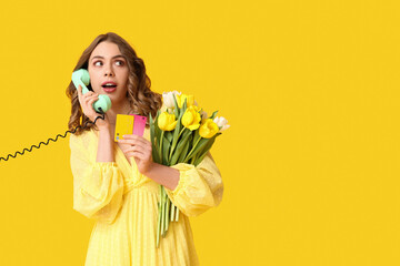 Young woman talking by retro telephone with bouquet of beautiful tulips and credit cards on yellow...