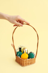 Female hand with wicker basket full of decorative cosmetics, Easter eggs and flowers on yellow background
