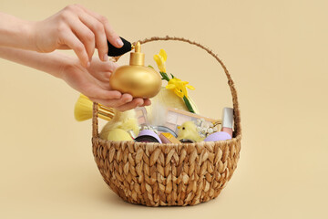 Female hands with wicker basket full of decorative cosmetics, baby chickens and Easter eggs on beige background