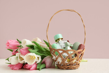 Wicker basket with decorative cosmetics, Easter eggs and bouquet of tulips on white table near pink wall