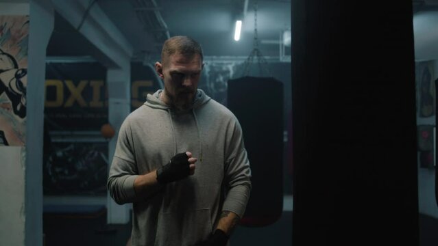 Athletic boxer stands in dark boxing gym and looks at camera. Male fighter in boxing bandages stretches, hits punching bag and prepares for training. Physical activity and intensive workout.
