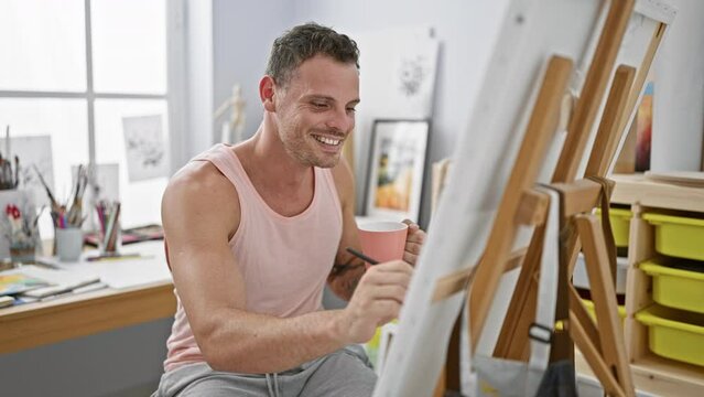 Smiling man paints in a bright studio while holding a coffee cup.