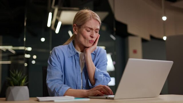 Disappointed young caucasian woman touching cheek and keeping eyes closed while working on wireless laptop in coworking space. Female employee feeling dental pain and discomfort at personal workplace.