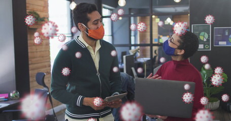 Image of covid 19 cells floating over man and woman in office wearing face masks - Powered by Adobe