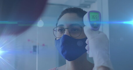 Image of glowing light trails over woman wearing face mask having temperature checked