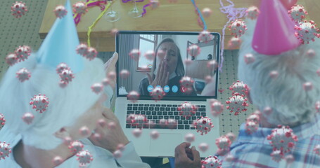 Image of covid 19 cells floating over senior couple in party hats using laptop on image call