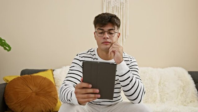 Confident young hispanic guy, positive expression glowing as he enjoys relaxing at home, resting comfortably on the sofa, using glasses to navigate online life on his touchpad in his cozy living room.