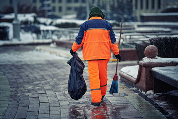 Municipal worker sweeps street, janitor with broom and dustpan working during snow storm in the evening. Man in uniform with broomstick and scoop sweep city street in winter season, cleanup sidewalk - obrazy, fototapety, plakaty