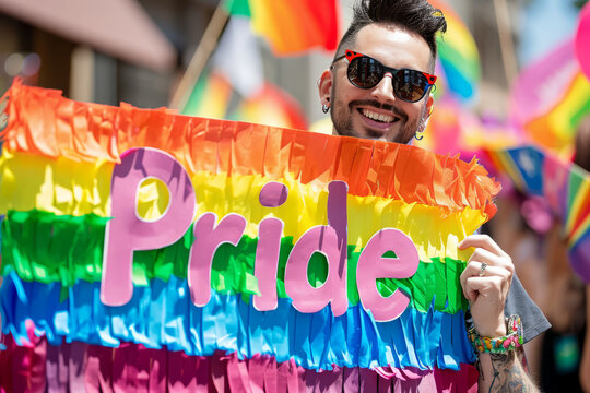 Happy gay man at a pride march holding a rainbow colored sign with written Pride text word at LGBTQ+ Gay Pride Parade