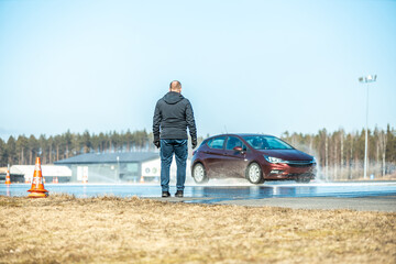 man observing a wet road test with a maroon car skidding on a water-covered track, testing vehicle safety and tire performance under controlled conditions
