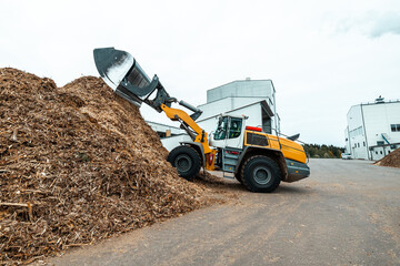 industrial wheel loader with raised bucket at a biomass power plant, handling wood chips against a backdrop of storage silos and blue sky, showcasing renewable energy resources