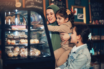 Smiling muslim mother wearing a hijab and her two young daughters looking at cakes in a cafe display case during a weekend activity together