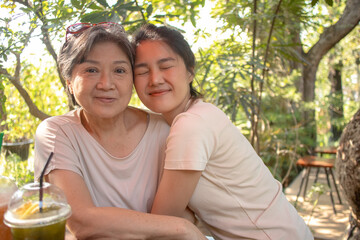 Asian Thai Chinese elder mother and daughter cuddling and take photo look at camera, drinking beverages at cafe garden, spend time together.