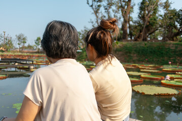 Backside view of asian Thai Chinese elder mother and daughter sitting on the bank by lotus flower lake local lagoon, both woman happy smiling, travel on vacation.