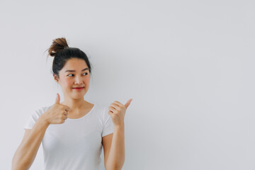 Asian Thai woman giving thumbs up, good recommend hand gesture, happy smiling looking at empty space, advertising standing isolated over white background wall.