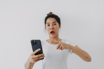 Asian Thai woman holding mobile phone with funny surprised and shocked face, while looking at camera, standing isolated over white background wall.
