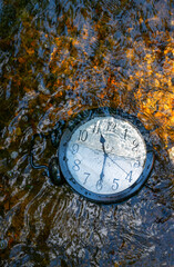 A large clock half submerged in a flowing stream of water with pebbles and ripples. Vertical image