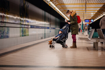 Mother and Child at Subway Station. A masked mother holding coffee pushes a stroller with her child at a subway station