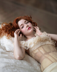 A girl with long red hair in a lace chemise with a corset lies on the bed