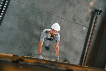 Going up on the ladder, top view. Young factory worker in grey uniform