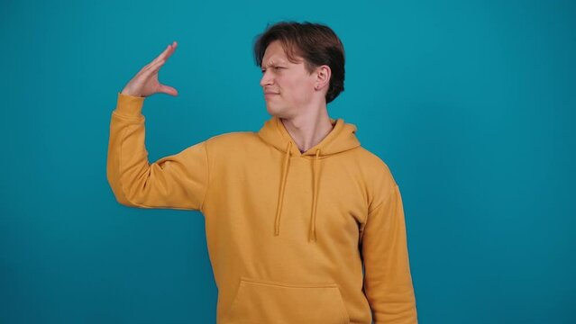 Young man shows bla-bla-bla gesture. Guy in yellow jacket imitates talking with hand, standing on blue background in studio. Babbling.