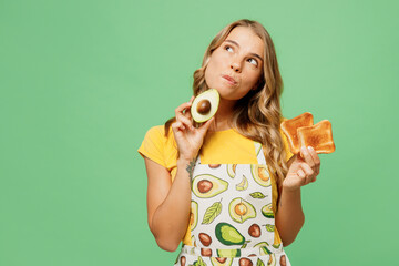 Young minded housewife housekeeper chef cook baker woman wear apron yellow t-shirt hold in hand avocado, bread toast look aside isolated on plain pastel green background studio. Cooking food concept.