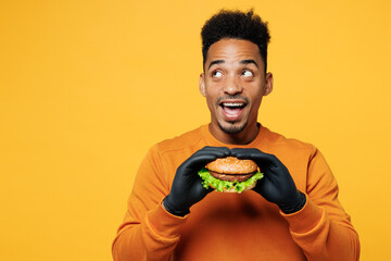 Young happy surprised shocked man wear orange sweatshirt casual clothes eat burger look aside on...