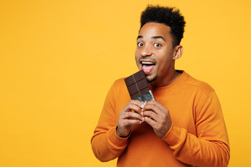 Young happy man wear orange sweatshirt casual clothes hold eat biting bar of chocolate isolated on plain yellow background studio portrait. Proper nutrition healthy fast food unhealthy choice concept. - obrazy, fototapety, plakaty