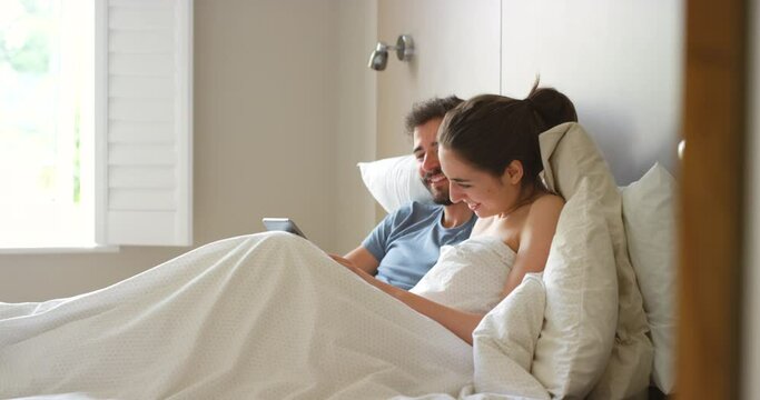 Happy couple, bed and laughing with tablet, touch and kiss while on vacation or relaxing at home. Man, woman and bonding by digital entertainment, online and laugh together at social media and jokes