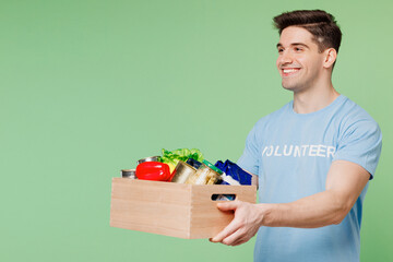 Young man wear blue t-shirt white title volunteer holding give box with vegetarian vegan food vegetables isolated on plain green background. Voluntary free work assistance help charity grace concept.