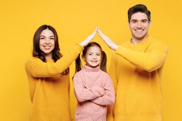Obraz premium Young fun parents mom dad with child girl 7-8 year old wear pink knitted sweater casual clothes hold folded hands above kid head like house roof isolated on plain yellow background Family day concept