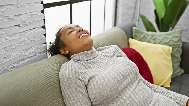A relaxed woman with curly hair enjoys tranquility on a sofa indoors at home, embodying contentment and leisure.