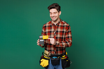 Young employee laborer man wear red shirt hold bank payment terminal to process acquire credit card isolated on plain green background. Instruments for renovation apartment room. Repair home concept.